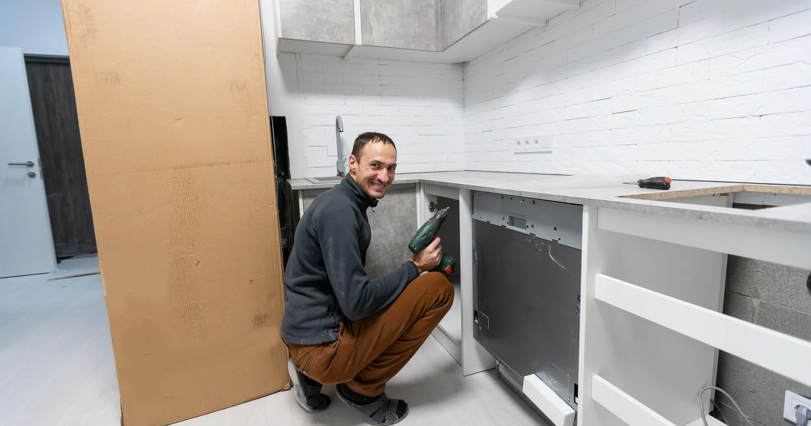 A handyman crouches in a bright white kitchen, drilling a base cabinet during installation.