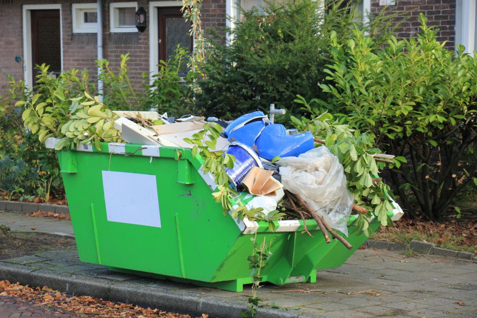 Green dumpster overflowing with debris and branches on a residential street in front of brick houses; leaves and bags spill over the sides.